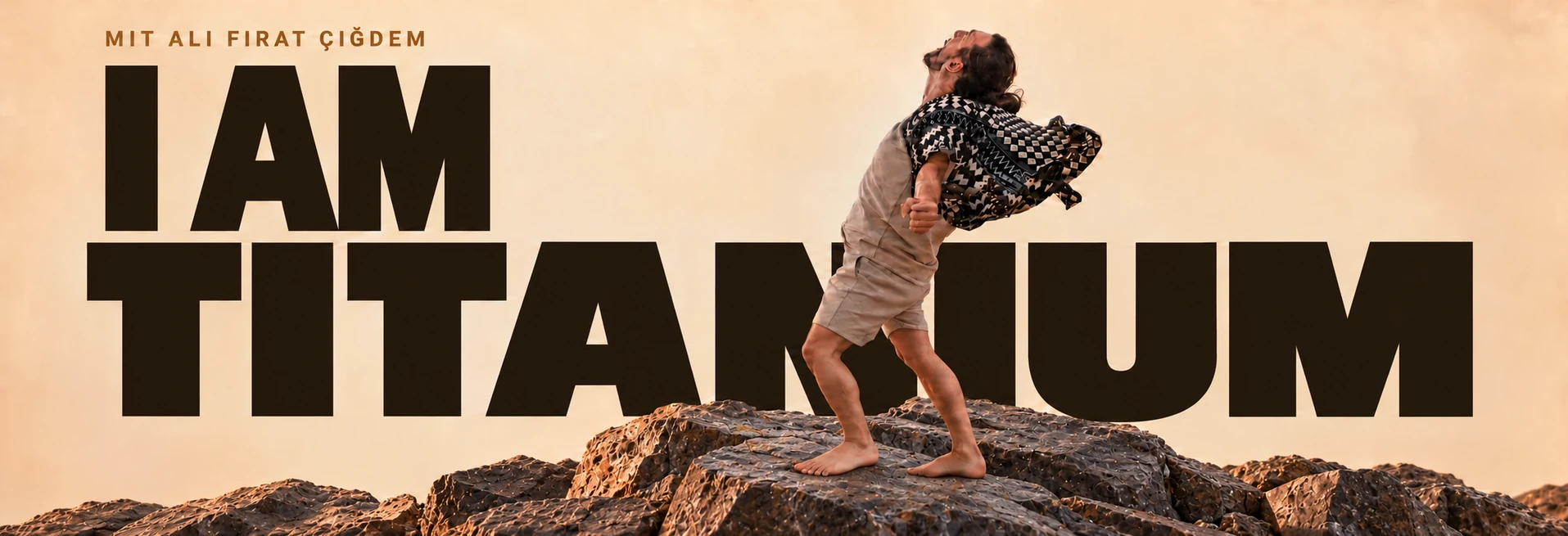I AM TITANIUM. Ali steht mit beiden vollständig sichtbaren Beinen und Füßen auf den Felsen, den Kopf zum Himmel gewandt. Warmer Sand, dunkle Schrift und das gemusterte Tuch im Wind.